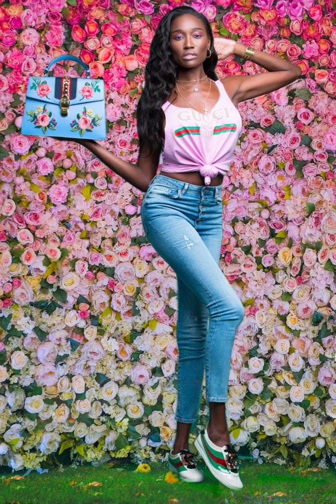 A woman holding a blue bag in front of a wall of flowers